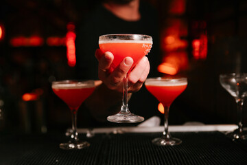 Bartender presenting a finished pink cocktail in a coupe glass with a sugar rim against a dark, atmospheric bar background