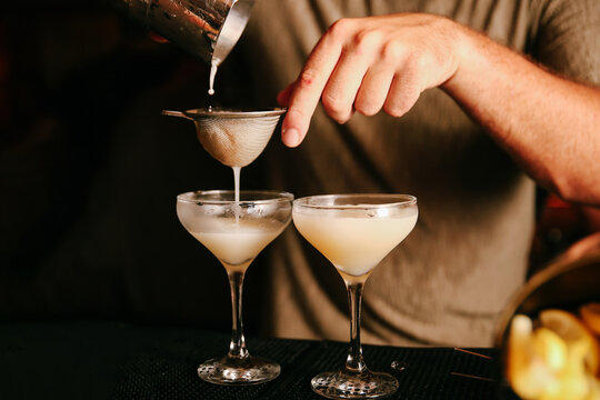 Close-up of a mixologist straining a clear cocktail into coupe glasses highlighting the delicate technique of mixology