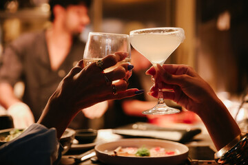 Close-up of female hands toasting with a cocktail and wine glass across a table in a dark atmospheric restaurant