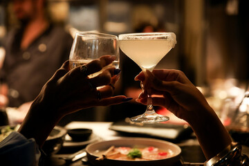 Close-up of female hands toasting with a cocktail and wine glass across a table in a dark atmospheric restaurant
