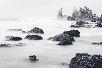 Minimalistic long exposure seascape with rocks emerging from soft misty water. Dramatic monochrome coastal landscape with atmospheric fog, textured stones and calm mood