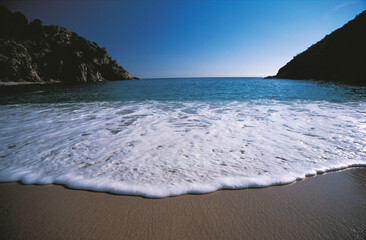 Clear blue sea with foamy wave gently reaching the sandy shore between rocky cliffs creating a peaceful coastal scene