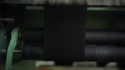 black webbing moving over rollers, textured woven strap advancing under tension control, rubberized drums and steady progression, dim workshop lighting, implied