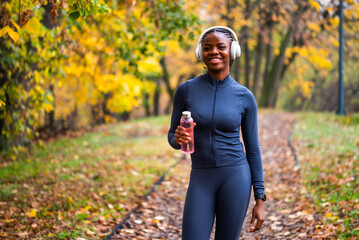 Obraz premium Black woman in athletic wear smiles while holding a water bottle, standing on a leaf-covered path surrounded by bright autumn foliage.