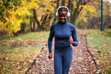 Happy woman jogs along a leaf-covered forest trail, surrounded by warm autumn colors and tall trees as she enjoys an outdoor run.