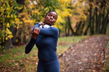 Young black woman is exercising in park in autumn. She is stretching her body before jogging.