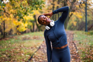 Young black woman is exercising in park in autumn. She is stretching her body before jogging.