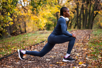 Young black woman is exercising in park in autumn. She is stretching her body before jogging.