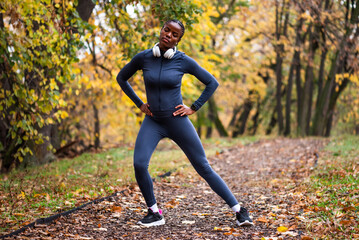 Young black woman is exercising in park in autumn. She is stretching her body before jogging.