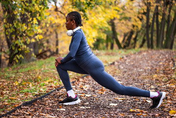 Young black woman is exercising in park in autumn. She is stretching her body before jogging.