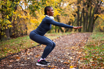 Young black woman is exercising in park in autumn. She is doing squats.