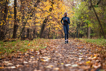 A smiling black woman runs on running trail of fallen leaves, with golden and green trees creating a serene, seasonal backdrop.