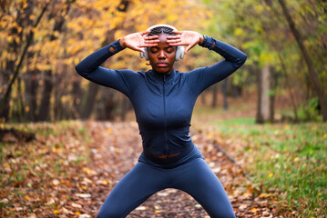 Young black woman is exercising Tai chi exercise in park in autumn. She is wearing wireless headphones.