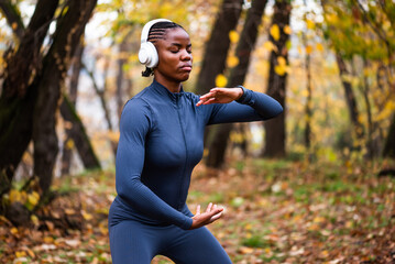 Young black woman is exercising Tai chi exercise in park in autumn. She is wearing wireless headphones.