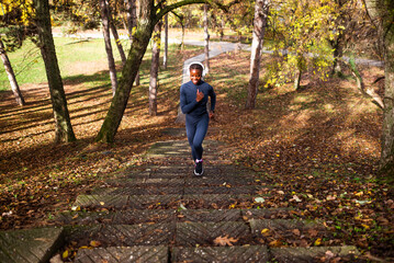 A woman jogs up a leaf-covered stone path in a park, wearing a navy athletic outfit and headphones, enjoying a sunny autumn day.
