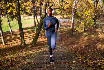 In a forested park with vibrant fall colors, a smiling runner in fitted sportswear climbs outdoor steps while listening to music.