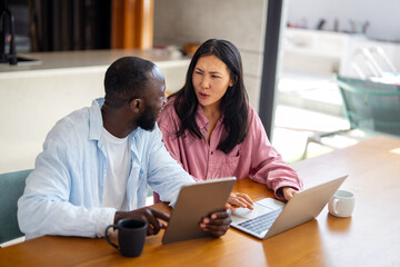 Multinational couple enjoying their morning routine, smiling and using laptop and tablet at home