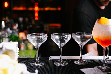 Row of coupe glasses prepared with salt rims and a large cocktail on a bar mat with a bartender ready to serve in the background