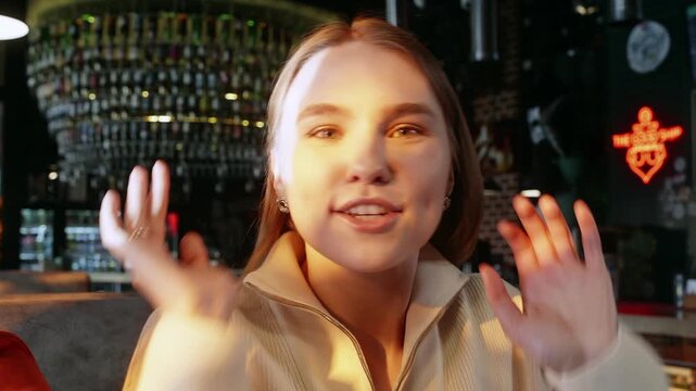 Young woman with light brown hair in beige zip jacket sits in cafe with glass decor, looks thoughtful, addresses boyfriend at side with warm expression, supports face with hand, warm ambient lighting