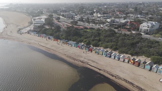 Aerial drone shot revealing the iconic colorful Brighton Bathing Boxes in Melbourne. Scenic coastline view of Dendy Street Beach, tourists, and suburbs on a hazy sunny day.