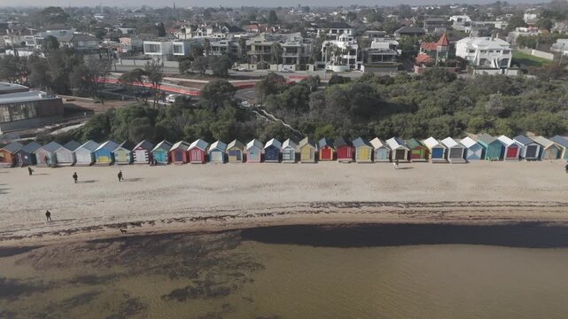 Aerial drone shot revealing the iconic colorful Brighton Bathing Boxes in Melbourne. Scenic coastline view of Dendy Street Beach, tourists, and suburbs on a hazy sunny day.