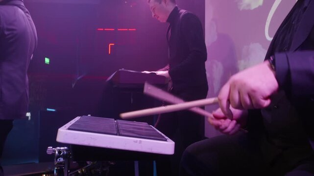 Dynamic shot of music band, male guitarist with glasses in dark suit, keyboardist in black turtleneck, drummer, purple and pink stage lighting, red accent lights, professional concert performance.