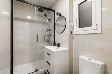 Modern bathroom interior with a glass shower, white sink, and toilet. Neutral tiles and natural light from a window create a clean, minimalist space.