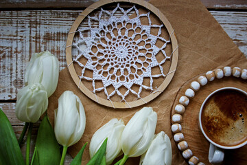 White crocheted doily in an embroidery hoop on kraft paper over a vintage white wooden background, paired with a cup of coffee and fresh white tulips, creating a bright spring craft and holiday mood.