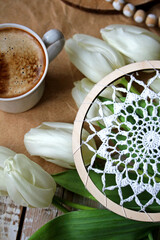 White crocheted doily in an embroidery hoop on kraft paper over a vintage white wooden background, paired with a cup of coffee and fresh white tulips, creating a bright spring craft and holiday mood.