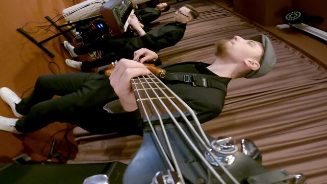 Vertical shot of young man stands on stage wooden floor playing dark green bass guitar, relaxed pose, amplifier and music equipment visible, music studio setting.