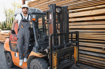 Warehouse worker standing on forklift near lumber stacks