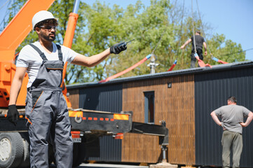 Construction worker supervising modular house transportation by crane and truck