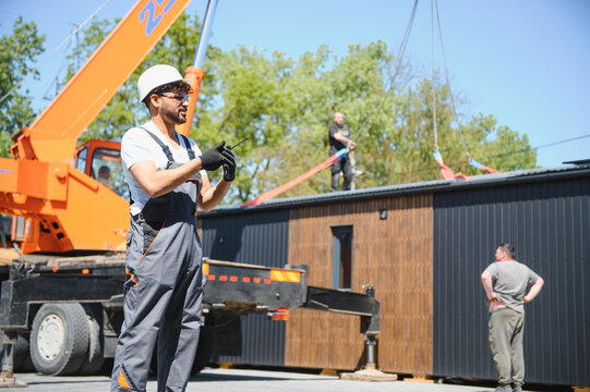 Construction worker using walkie talkie while lifting modular house with crane