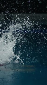 Focused swimmer in red cap gliding through dark indoor pool water, creating powerful splashes and ripples while practicing intense lap swimming technique