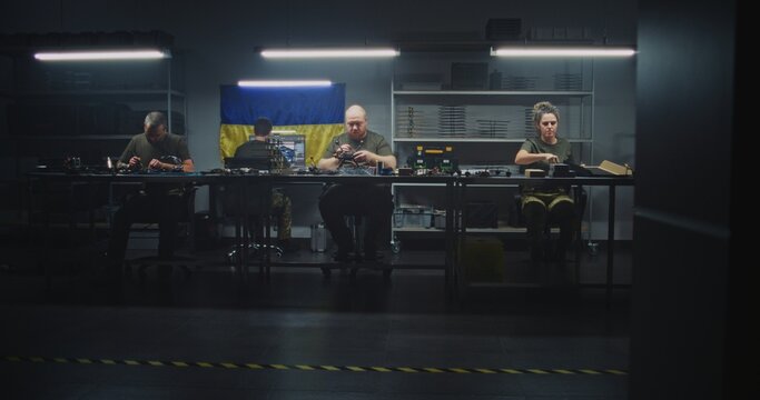 Male Worker in Camouflage Uniform Moves Equipment Cases Through Drone Facility, While Team Works on FPV Drone Assembly Under Ukrainian Flag. Concept of Logistics and Teamwork in Defense Tech.
