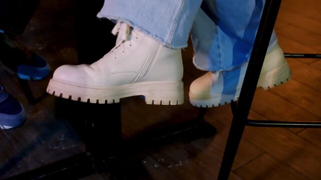 Close up of womans foot in white lace-up boots tapping in sync with music, casual frayed jeans, sitting on black bar stool, friendly atmosphere, warm light, relaxed pose, wood texture.