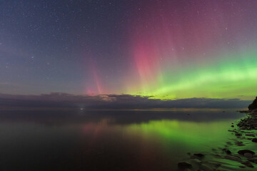 Stunning green and pink Aurora Borealis reflecting in calm Baltic Sea water near rocky Estonian coastline under starry night sky