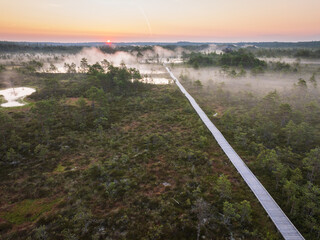 Aerial view of a wooden boardwalk winding through a misty peat bog landscape during a colorful sunrise