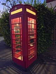 Obraz premium A classic red British telephone booth (kiosk) illuminated from within, standing on a dark pavement next to a dense green hedge and a brick wall, likely taken at twilight or night.
