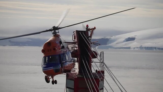 Bright orange helicopter lifting off from a ship's helipad in a polar seascape, windsock and railings visible, as it climbs over icy waters beneath an overcast sky.