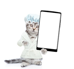 Cute kitten wearing shower cap and bathrobe standing on hind legs showing big smartphone with white blank screen in it paw. Isolated on white background