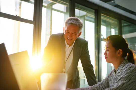 Senior businessman mentoring young employee in modern office, sunlight streaming through window, professional guidance and teamwork create positive work environment and career growth opportunities