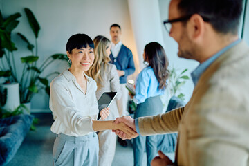In a corporate office, a business executive is seen smiling and shaking hands to seal a deal, with a group of diverse colleagues in the background