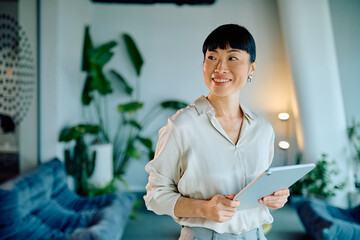 Confident Asian businesswoman holding a digital tablet with a happy expression, standing in a contemporary office setting