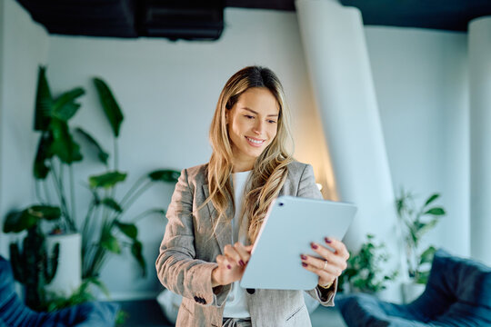 Smiling young businesswoman using a digital tablet for work, standing in a contemporary office space with plants