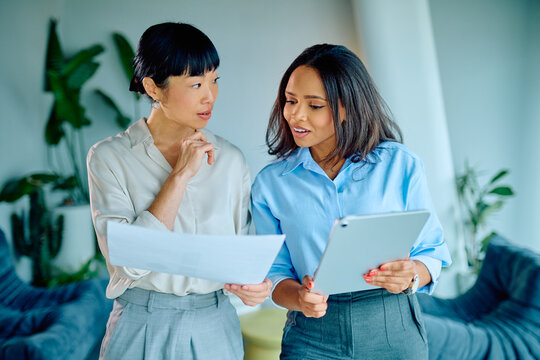 Two diverse businesswomen reviewing papers and a digital tablet, sharing ideas and working together in a contemporary office setting