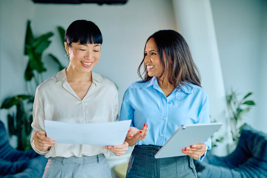 Two smiling businesswomen, one Asian and one Black, collaborating, discussing documents and digital tablet content while working