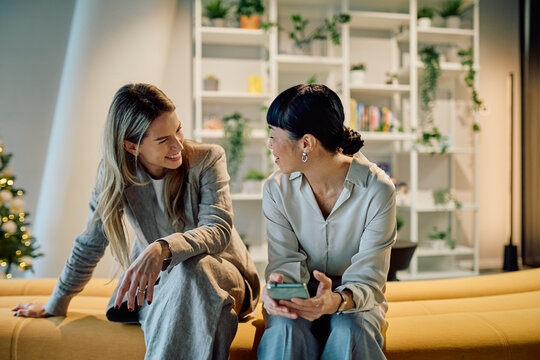 Two diverse businesswomen colleagues having a friendly conversation and smiling, sitting on a yellow sofa in a modern office environment