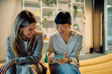 Two diverse businesswomen laughing and sharing content on a smartphone during a break, depicting connection, friendship, and teamwork