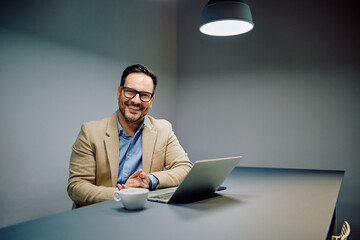 Cheerful man in business attire, wearing glasses, sitting at a modern workspace with a laptop and coffee mug, looking pleased as he poses for the camera